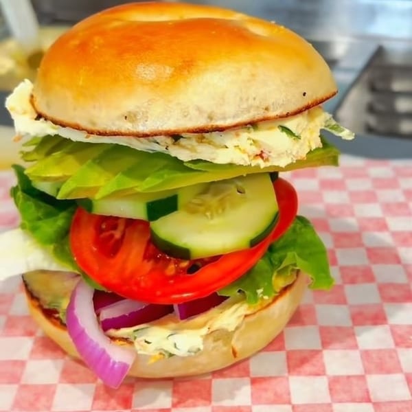 A bagel sandwich with egg salad, lettuce, tomato, cucumber, and red onion on red and white checkered paper.