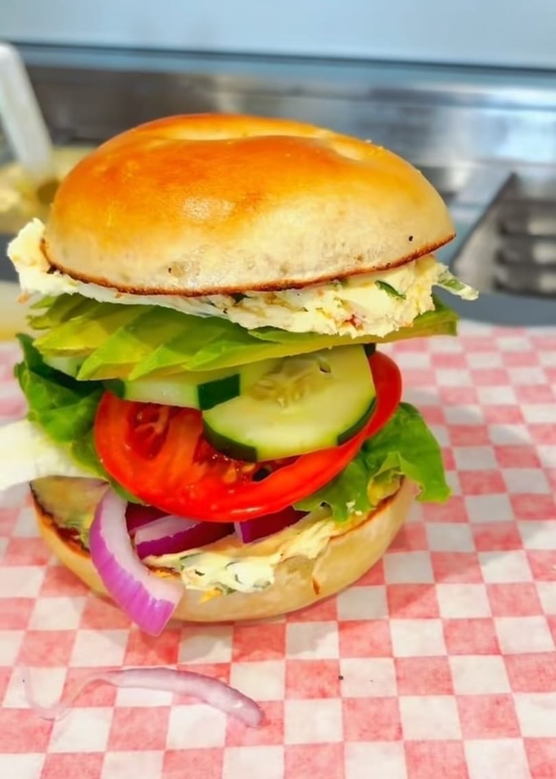 A bagel sandwich with egg salad, lettuce, tomato, cucumber, and red onion on red and white checkered paper.