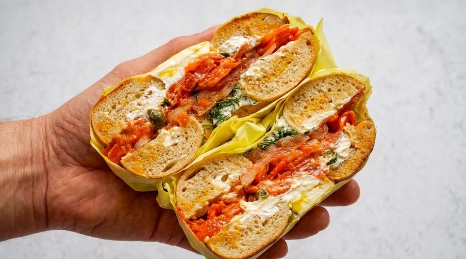 A hand holds a bagel sandwich, cut in half, filled with salmon, cream cheese, and dill.