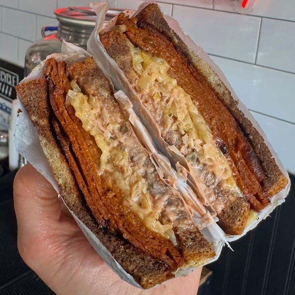 Hand holding a sliced sandwich with dark bread, meat, cheese, and sauce, wrapped in paper.