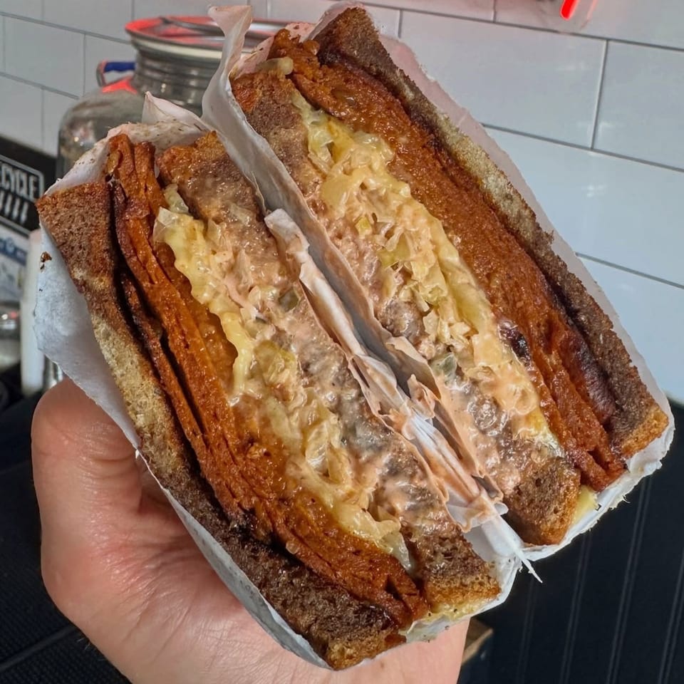 Hand holding a sliced sandwich with dark bread, meat, cheese, and sauce, wrapped in paper.