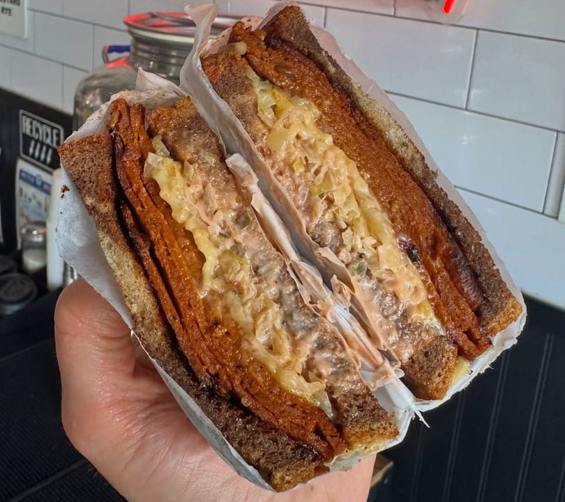 Hand holding a sliced sandwich with dark bread, meat, cheese, and sauce, wrapped in paper.