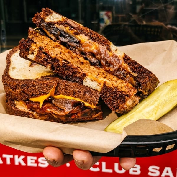 Hand holding a basket with a Latkes Club Sandwich, cut in half, and a pickle spear.