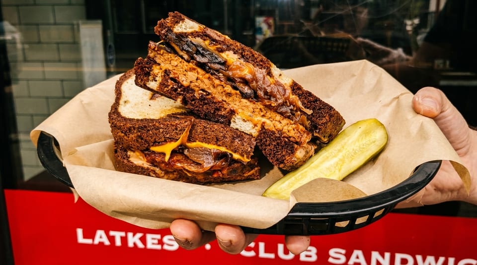 A hand holds a basket with a Latkes Club Sandwich and a pickle spear.