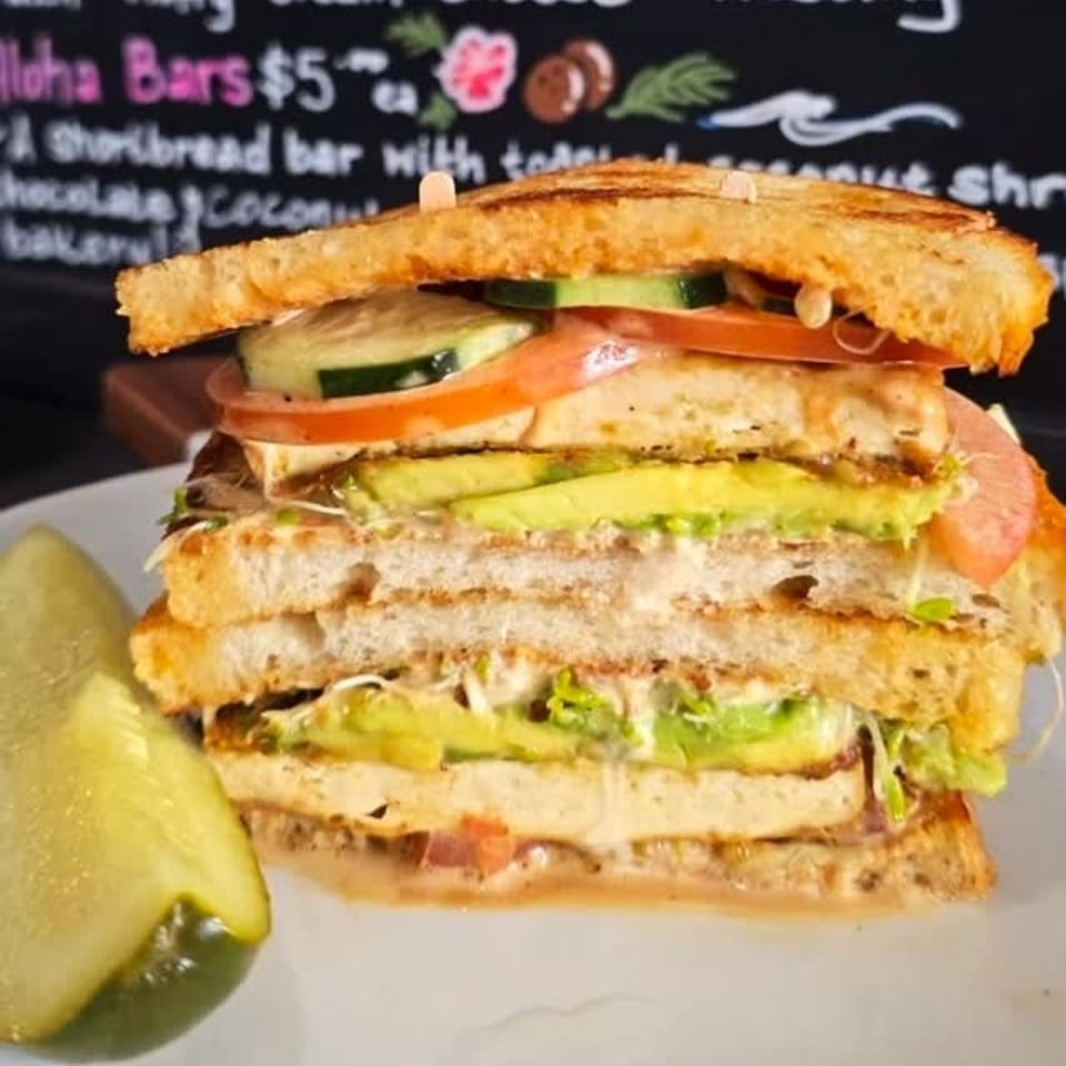 A tall, layered sandwich with avocado, tomato, cucumber, and sprouts, served with a pickle spear.