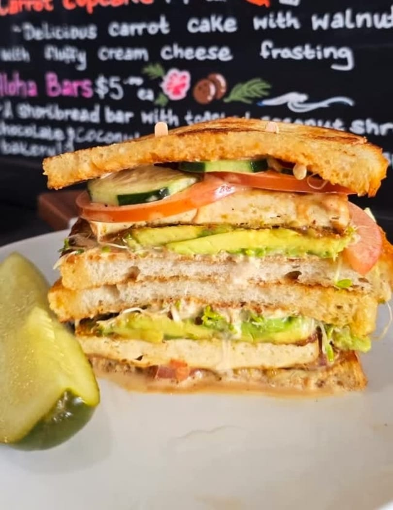 A tall, layered sandwich with avocado, tomato, cucumber, and sprouts, served with a pickle spear.