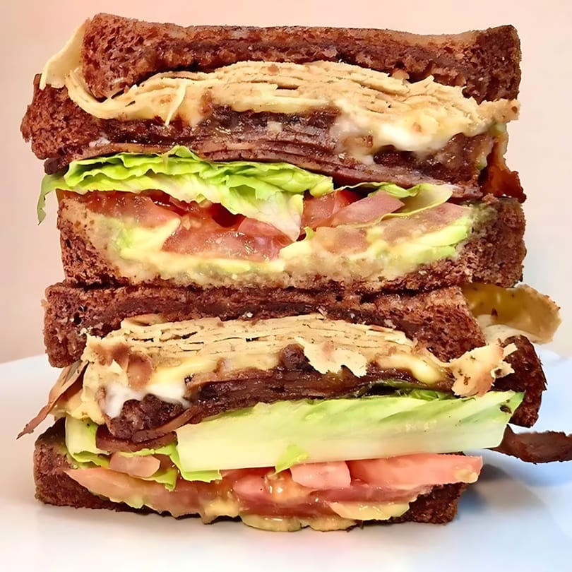 A towering double-decker sandwich with dark bread, meat, cheese, lettuce, tomato, and avocado.