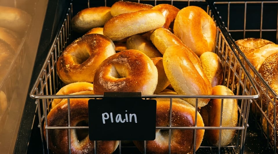 A wire basket filled with golden-brown plain bagels, with a small "Plain" sign in front.