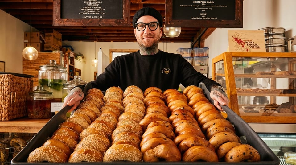 A tattooed man in a beanie and glasses holds a large tray of freshly baked bagels.