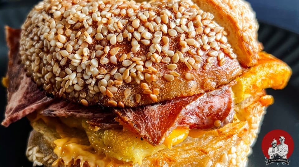 Close-up of a sesame seed bagel sandwich with pastrami, melted cheese, and egg.